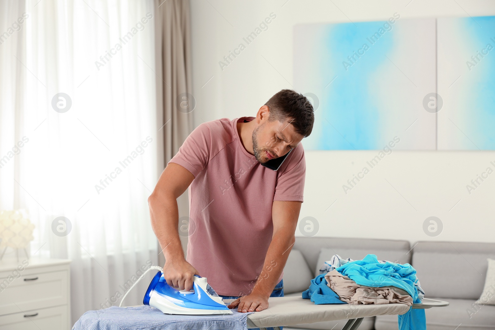 Photo of Handsome man ironing clean laundry while talking smartphone at home