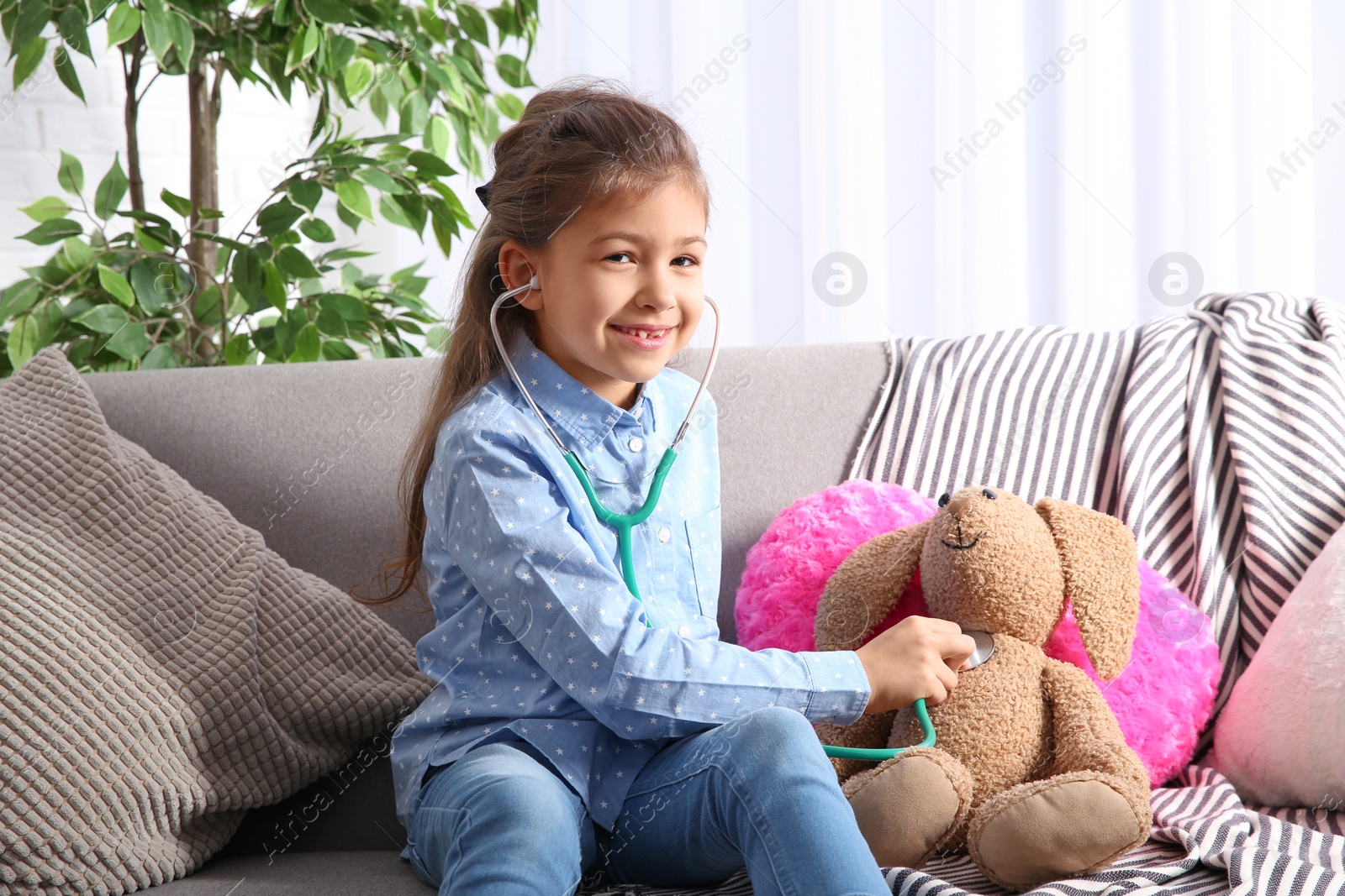 Photo of Cute child imagining herself doctor while playing with stethoscope on sofa in living room