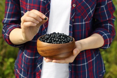 Photo of Woman putting bilberry into wooden bowl on blurred background, closeup