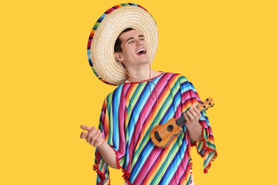 Photo of Young man in Mexican sombrero hat and poncho playing ukulele on yellow background