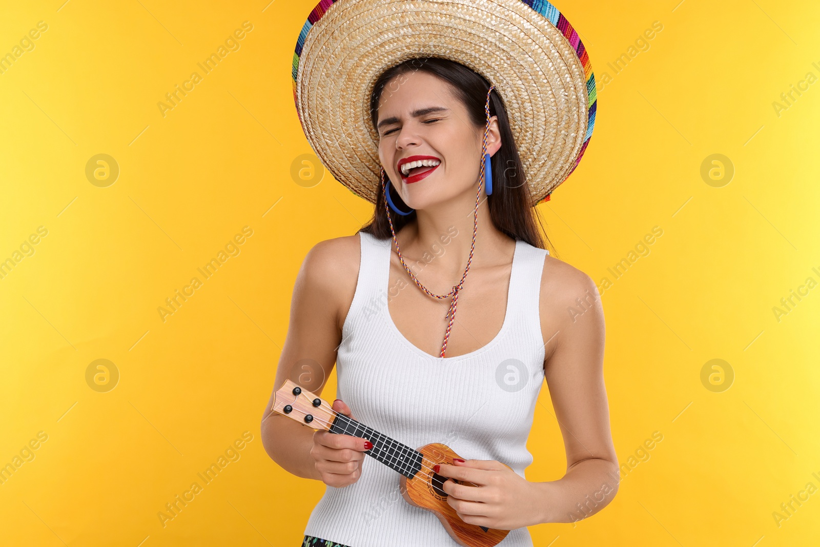 Photo of Young woman in Mexican sombrero hat playing ukulele on yellow background