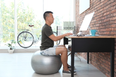 Photo of Young man sitting on fitness ball and using computer in office. Workplace fitness