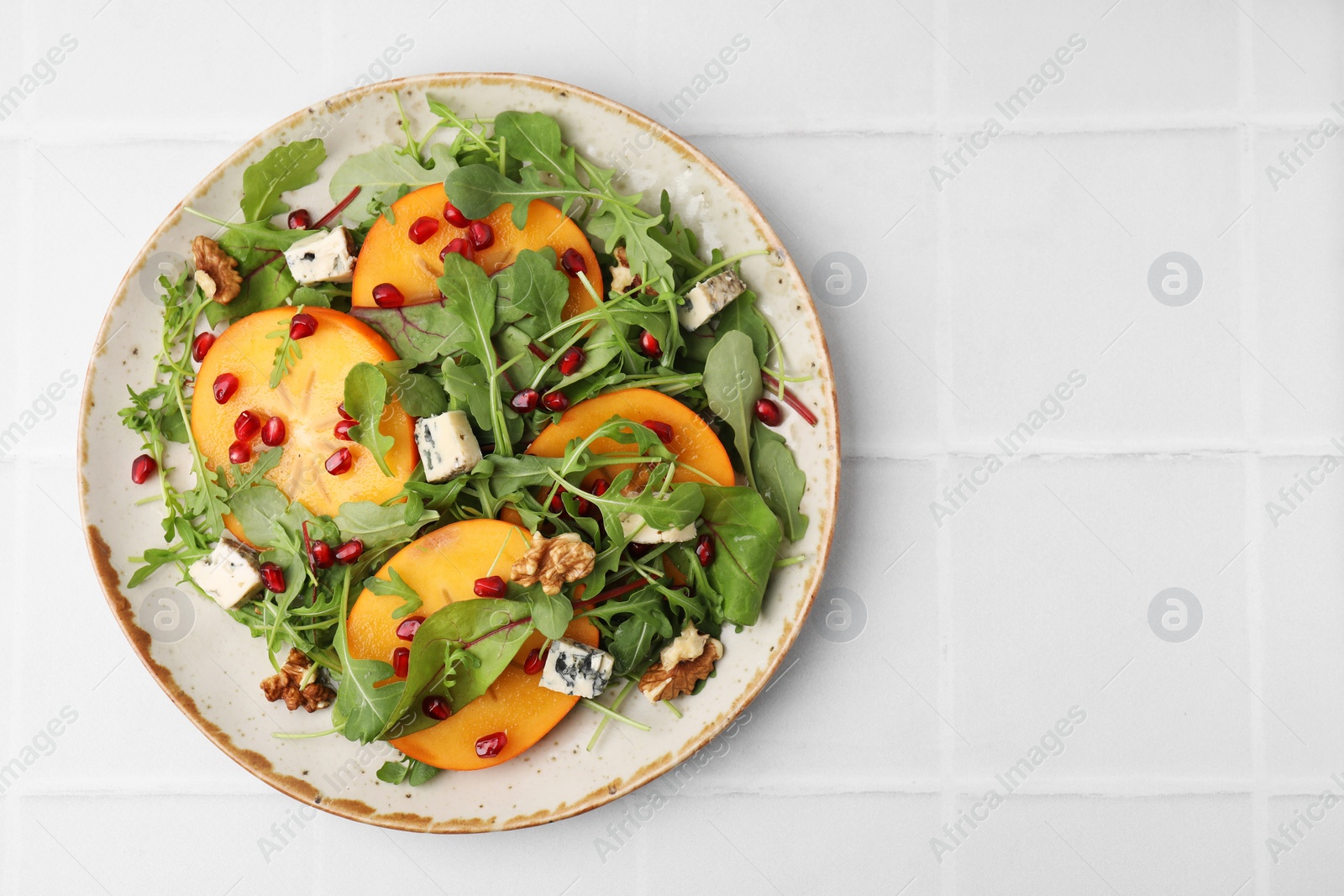 Photo of Tasty salad with persimmon, blue cheese, pomegranate and walnuts served on white tiled table, top view. Space for text