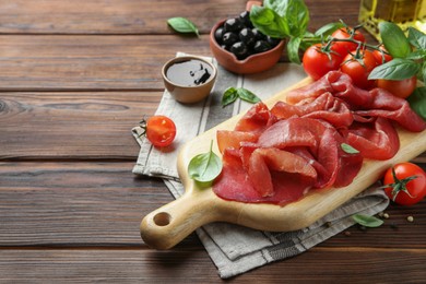 Photo of Board with delicious bresaola served with tomato and basil leaves on wooden table. Space for text