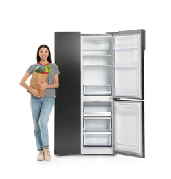 Young woman with bag of groceries near open empty refrigerator on white background