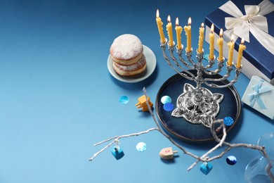 Photo of Hanukkah celebration. Menorah with burning candles, dreidels, donuts and gift boxes on light blue table, above view. Space for text