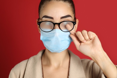 Photo of Woman wiping foggy glasses caused by wearing medical mask on red background