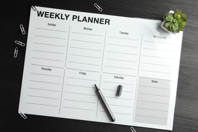 Photo of Timetable. Weekly planner, felt pen, houseplant and paper clips on black wooden table, top view