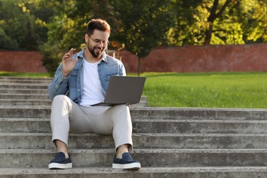 Photo of Handsome young man with laptop sitting on concrete stairs outdoors. Space for text