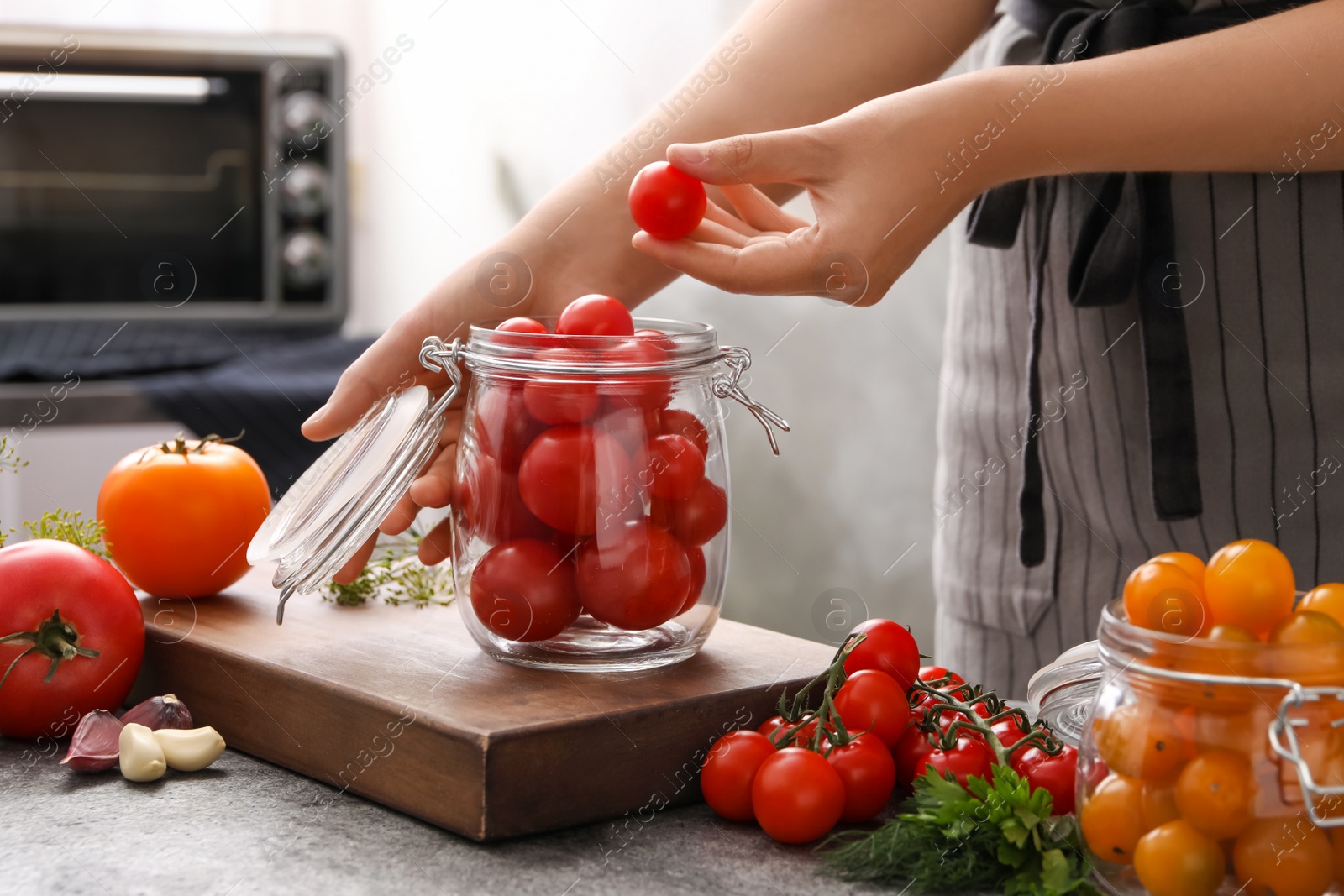 Photo of Woman putting tomatoes into glass jar at grey kitchen table, closeup. Pickling vegetables
