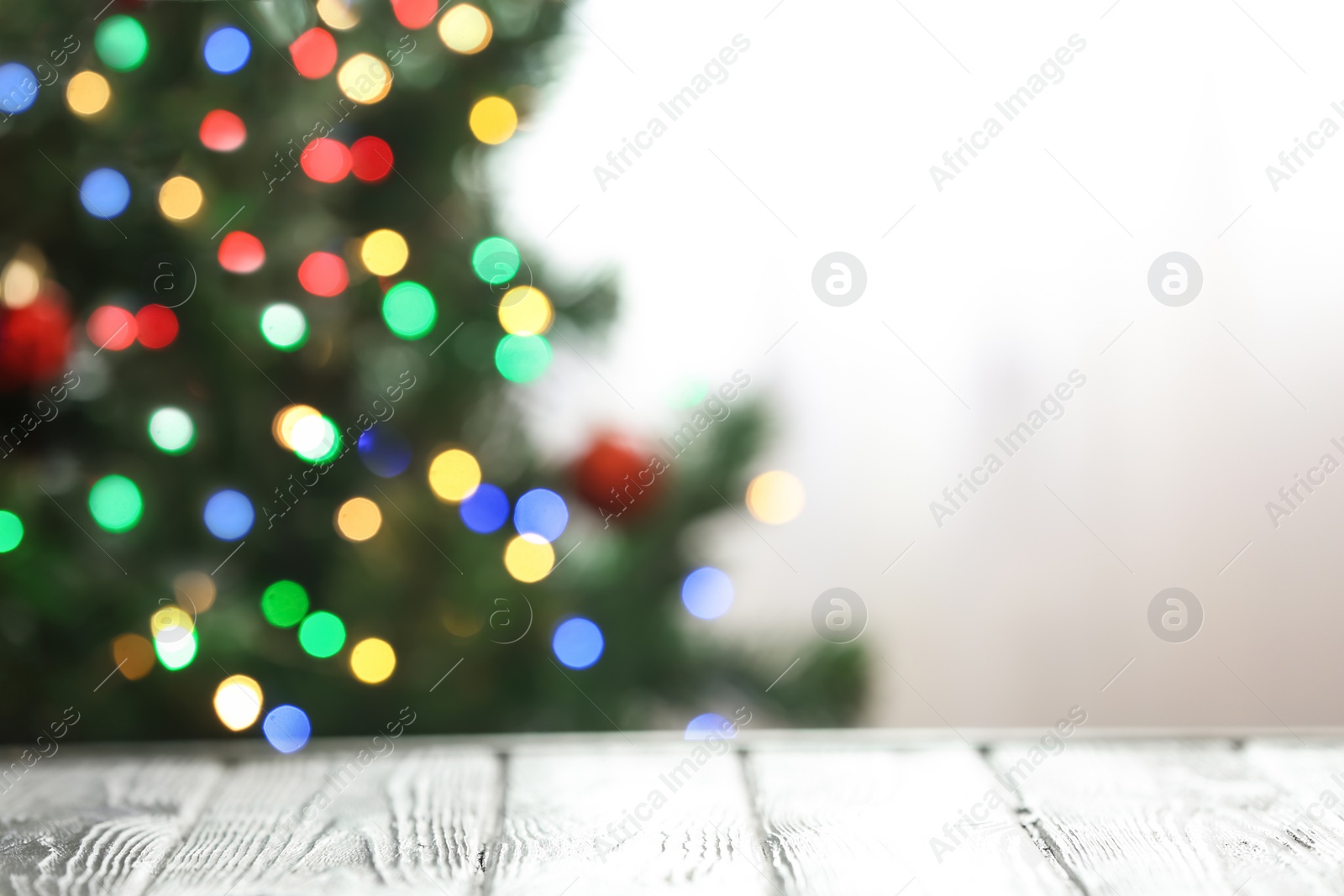 Photo of Blurred view of fir tree with glowing Christmas lights near table indoors. Festive mood