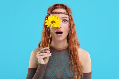 Photo of Surprised young hippie woman covering eye with sunflower on light blue background