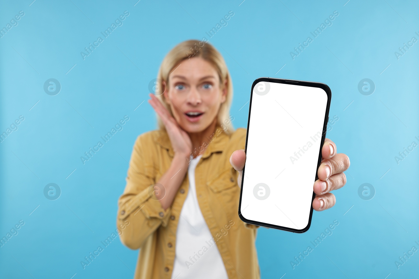 Photo of Surprised woman holding smartphone with blank screen on light blue background, selective focus