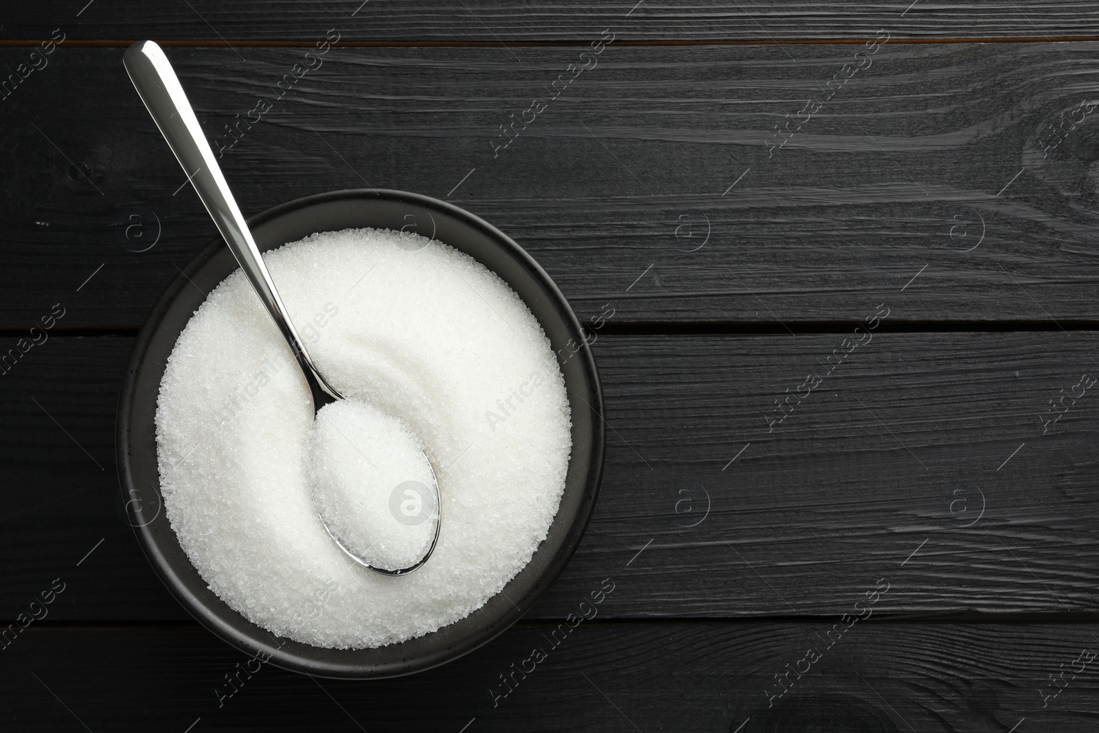 Photo of Granulated sugar in bowl and spoon on black wooden table, top view. Space for text
