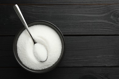 Photo of Granulated sugar in bowl and spoon on black wooden table, top view. Space for text