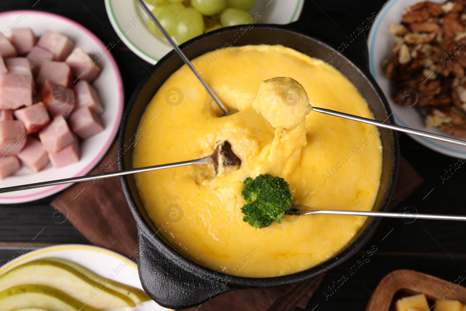 Photo of Dipping different products into fondue pot with melted cheese on black wooden table, flat lay