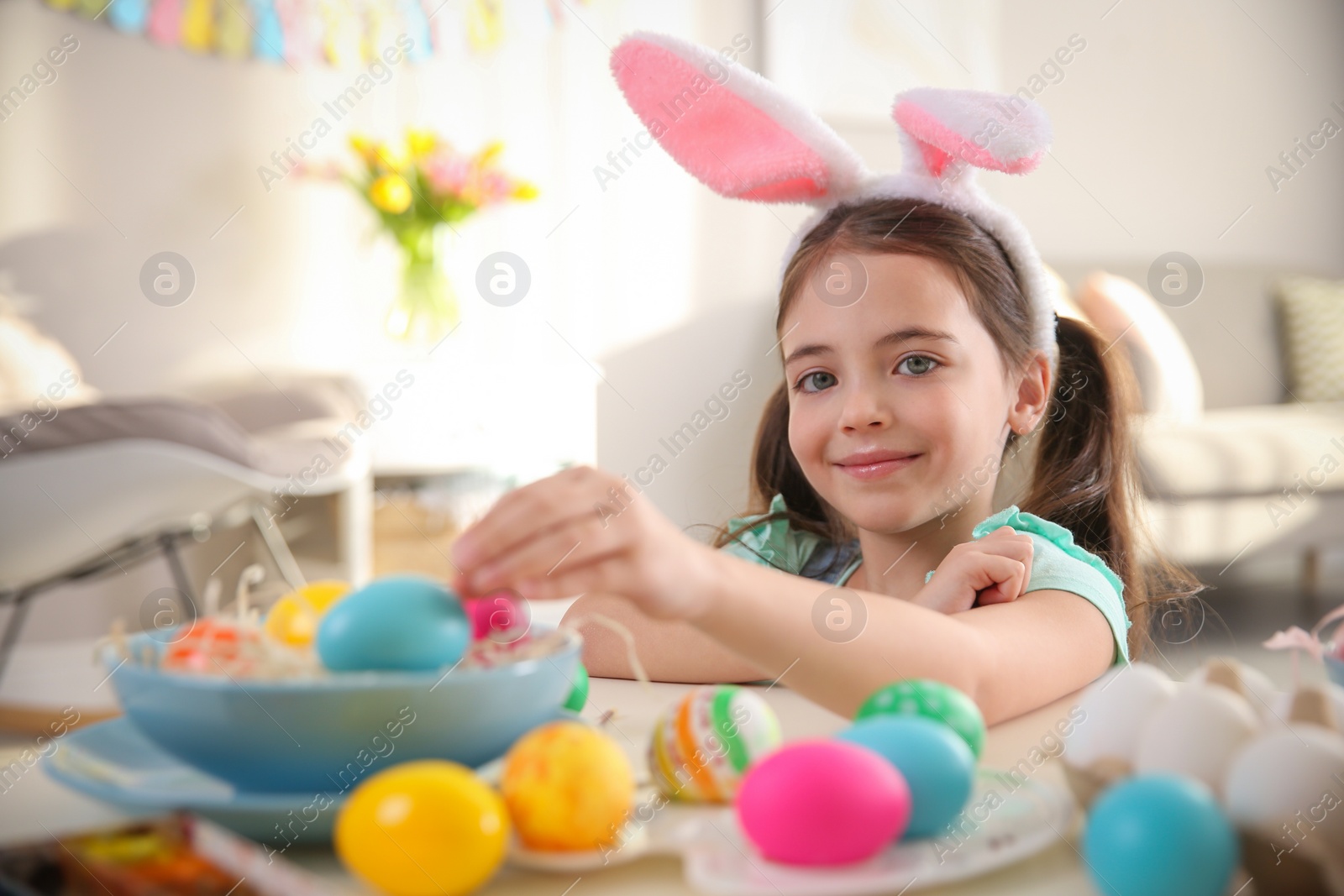 Photo of Cute little girl in bunny ears headband painting Easter eggs at table indoors