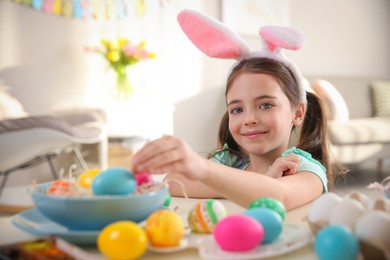 Cute little girl in bunny ears headband painting Easter eggs at table indoors