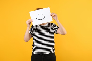 Photo of Woman hiding behind sheet of paper with happy face on yellow background