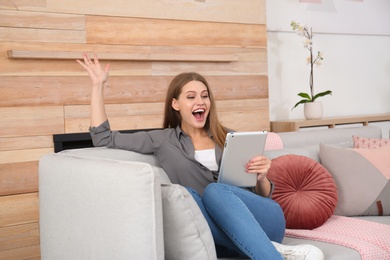 Photo of Emotional young woman with tablet celebrating victory on sofa at home