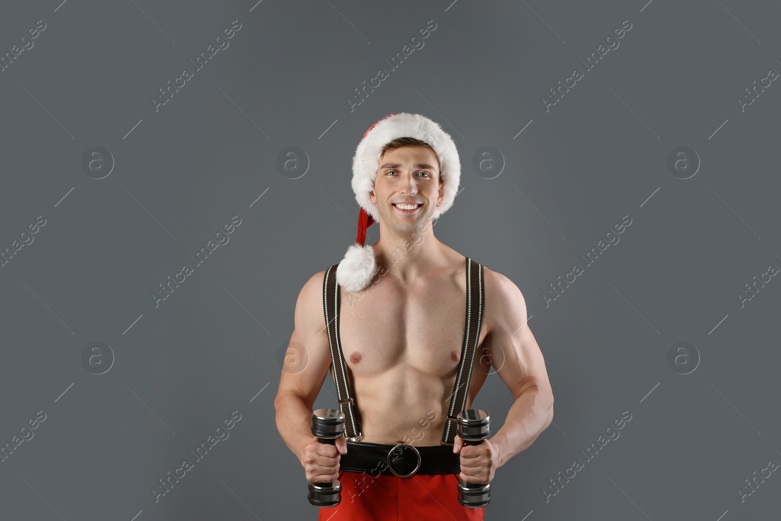 Photo of Young muscular man in Santa hat with dumbbells on gray background