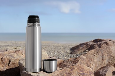 Photo of Metallic thermos and cap on stone near sea, space for text