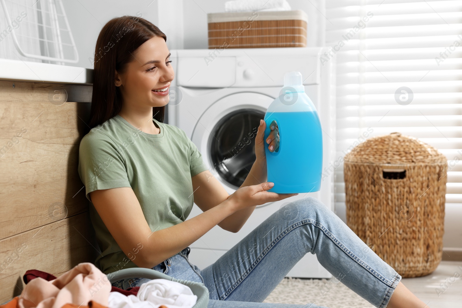 Photo of Woman sitting near washing machine and holding fabric softener in bathroom
