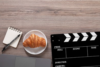 Photo of Movie clapper, croissant, notebook and pen on wooden table, flat lay. Space for text
