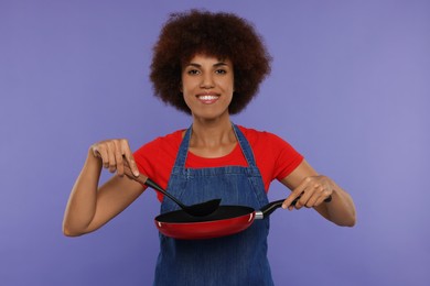 Photo of Happy young woman in apron holding ladle and frying pan on purple background
