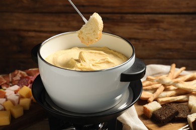 Dipping piece of bread into fondue pot with melted cheese at table with snacks, closeup