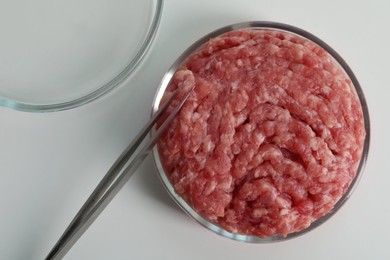 Petri dish with raw minced cultured meat and tweezers on white table, flat lay