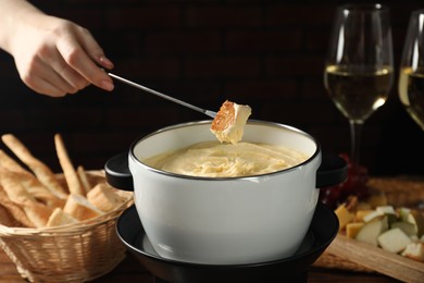 Photo of Woman dipping piece of bread into fondue pot with melted cheese at table, closeup