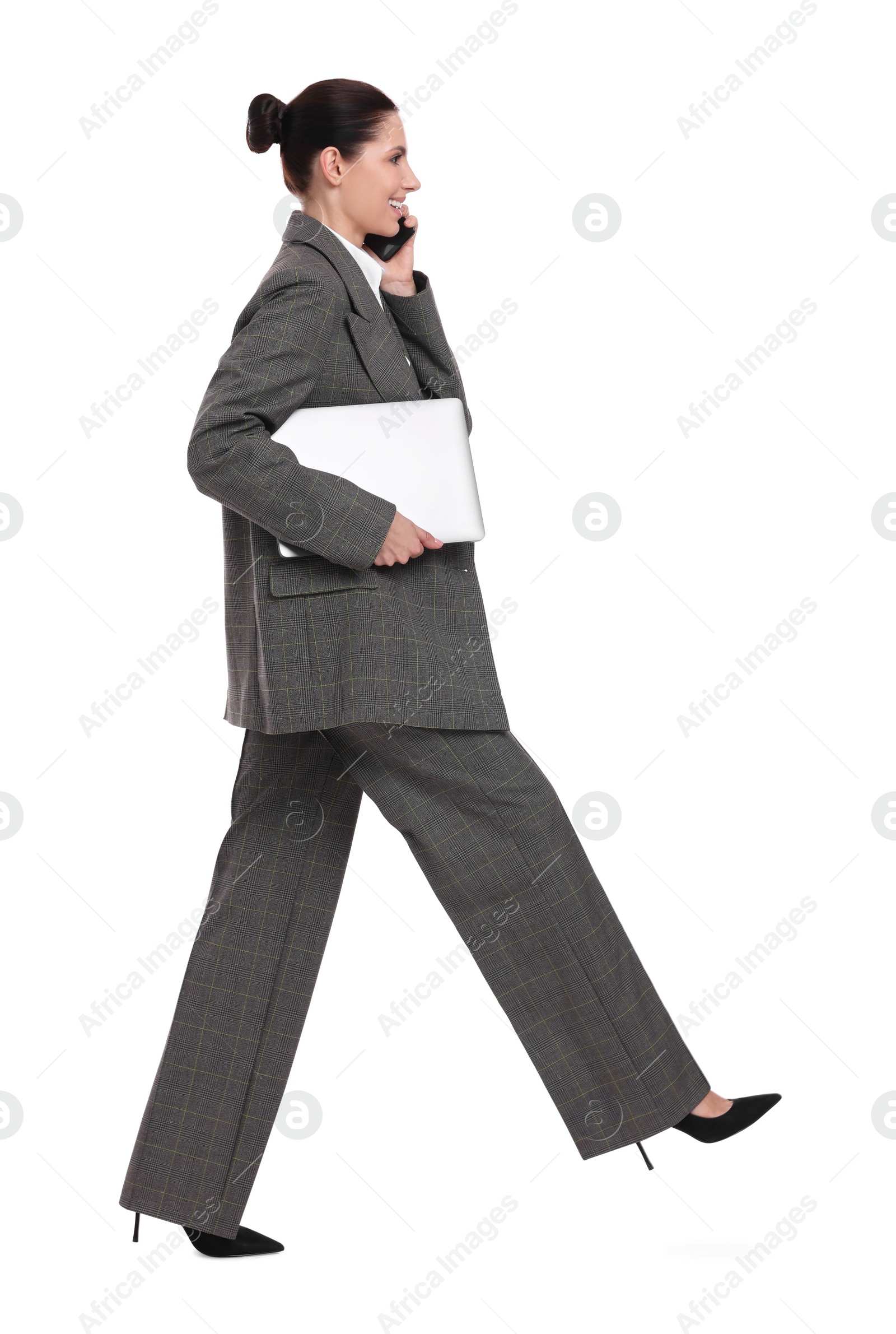 Photo of Beautiful happy businesswoman with laptop talking on smartphone while walking against white background