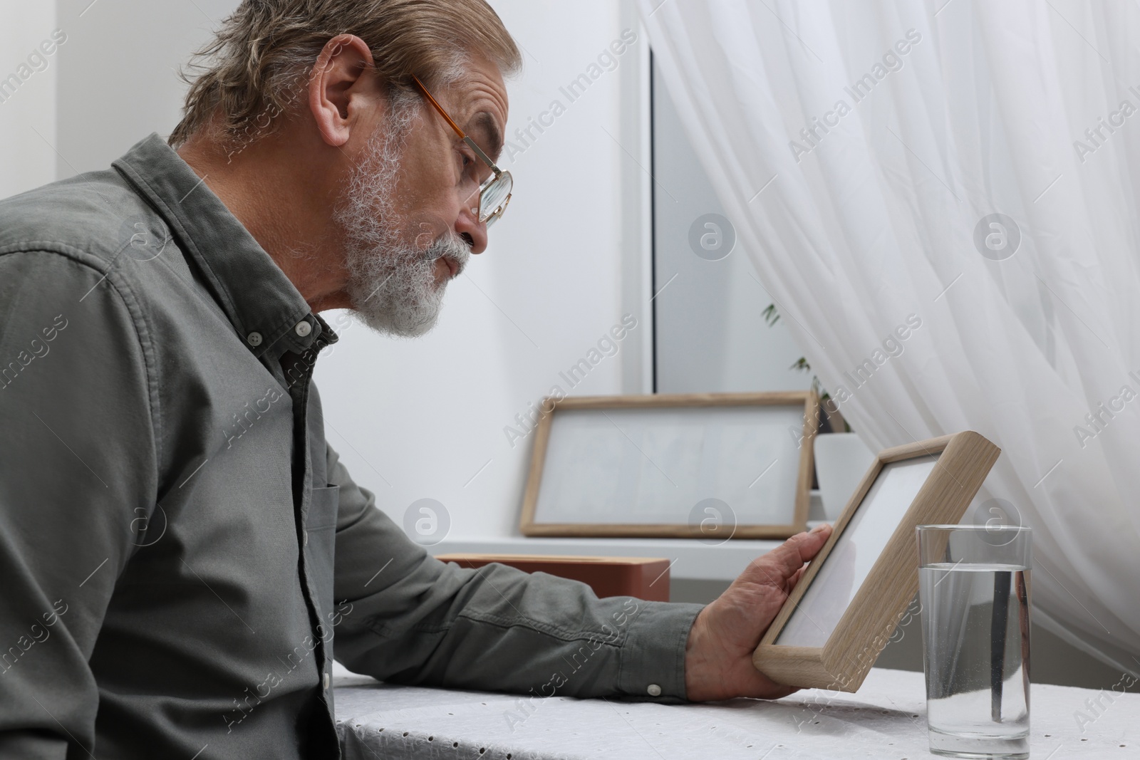 Photo of Upset senior man with photo frame at table in room. Loneliness concept