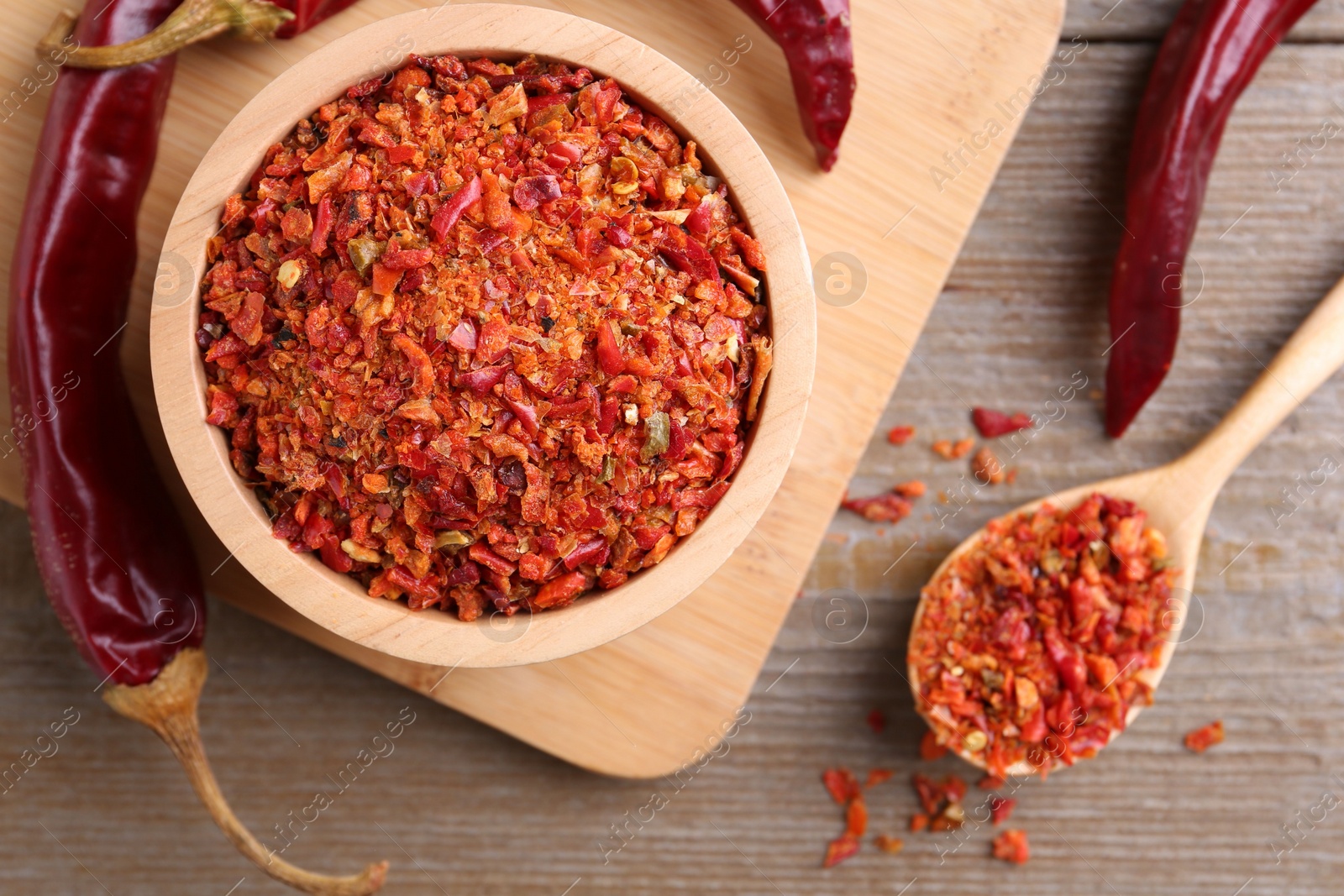 Photo of Aromatic spice. Red chili pepper flakes in bowl, spoon and pods on wooden table, flat lay