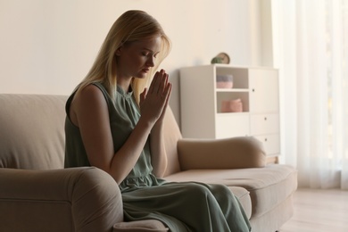 Religious young woman praying on sofa at home. Space for text