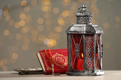 Photo of Arabic lantern, Quran and misbaha on table against blurred lights