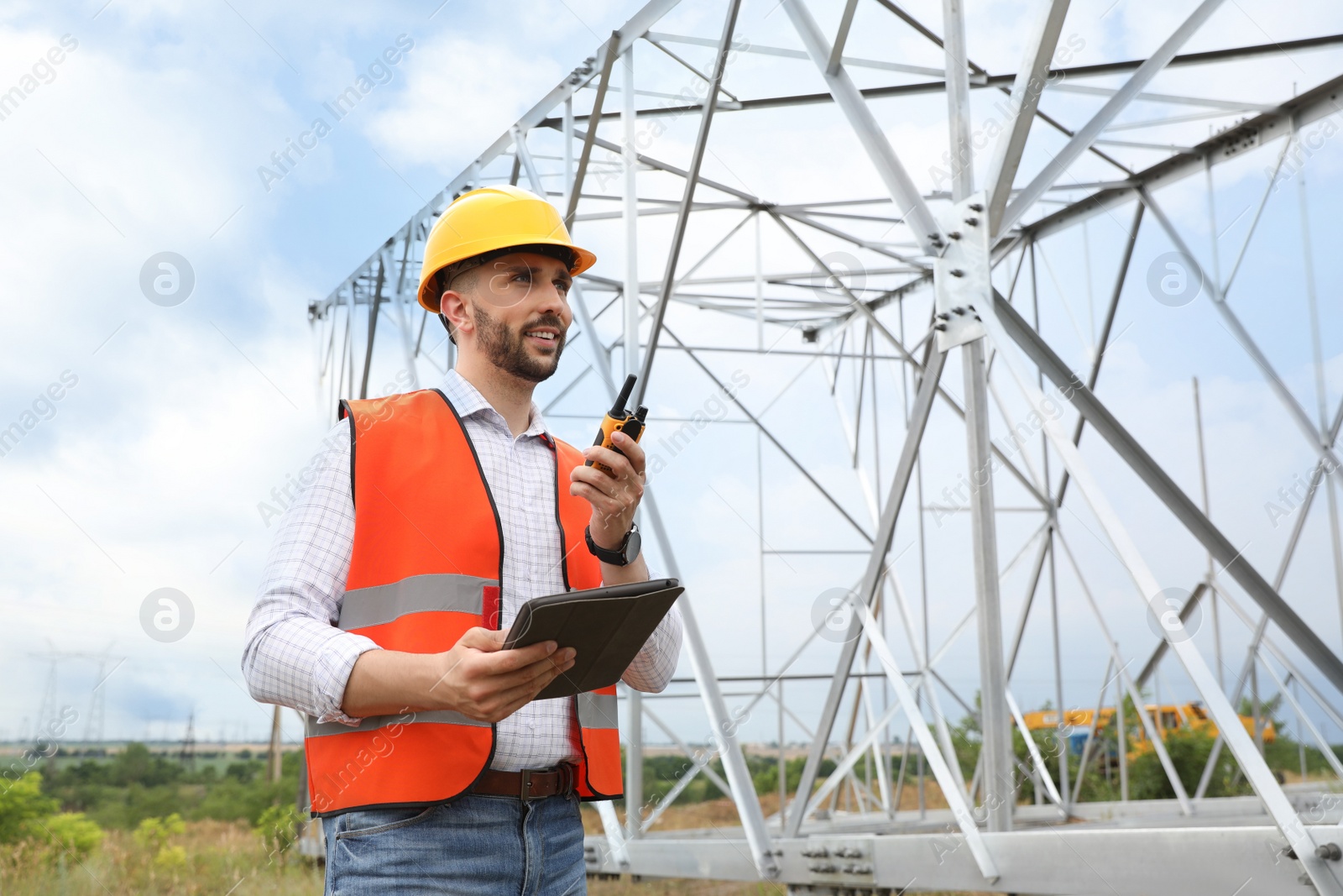 Photo of Professional engineer with tablet and walkie talkie near high voltage tower construction outdoors. Installation of electrical substation