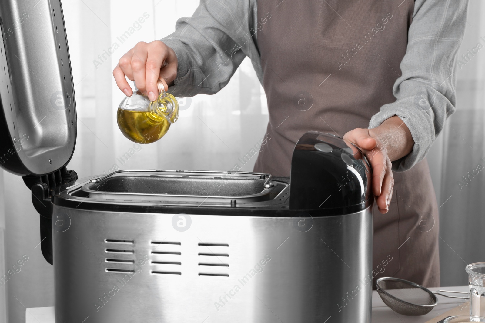 Photo of Woman pouring oil into breadmaker at white wooden table indoors, closeup