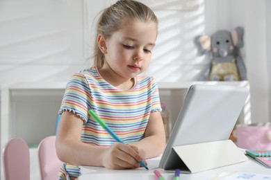 Photo of Little girl learning English with tablet indoors at online lesson