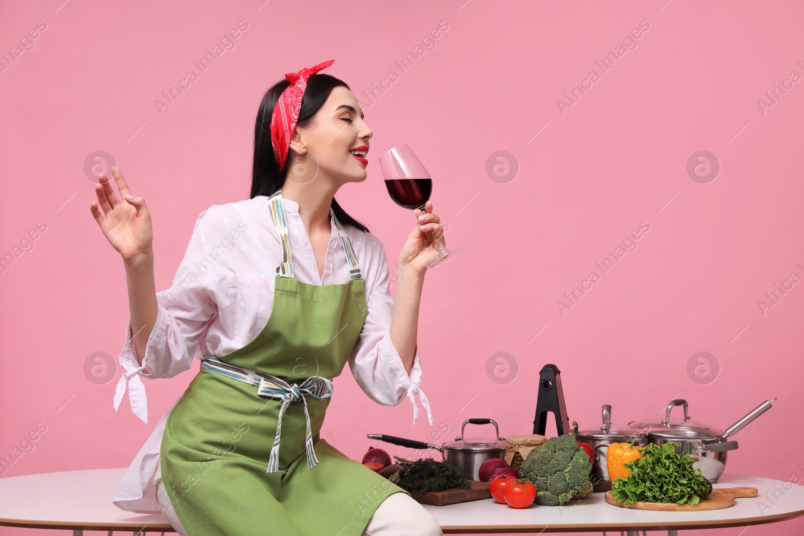 Photo of Young housewife with glass of wine, vegetables and different utensils on pink background. Space for text