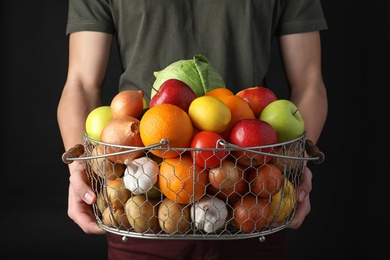 Photo of Man holding basket full of fresh  vegetables and fruits against black background, closeup