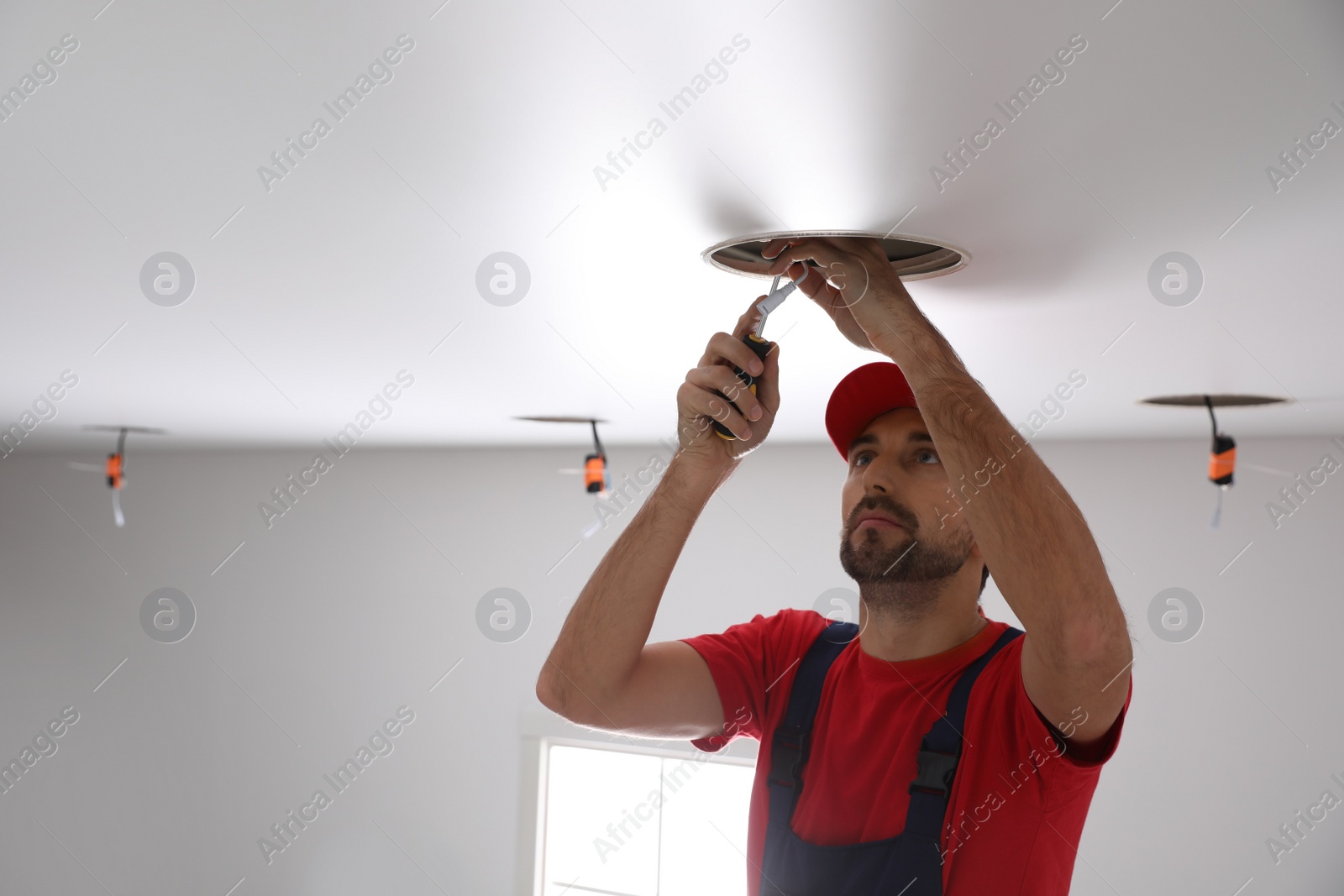 Photo of Worker installing lamp on stretch ceiling indoors