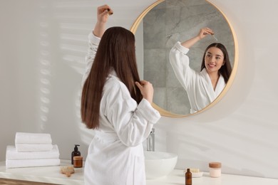 Photo of Happy young woman applying essential oil onto hair roots near mirror in bathroom