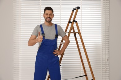 Worker in uniform and stepladder near horizontal window blinds indoors