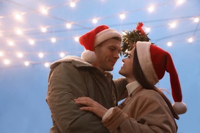 Happy couple in Santa hats standing under mistletoe bunch outdoors