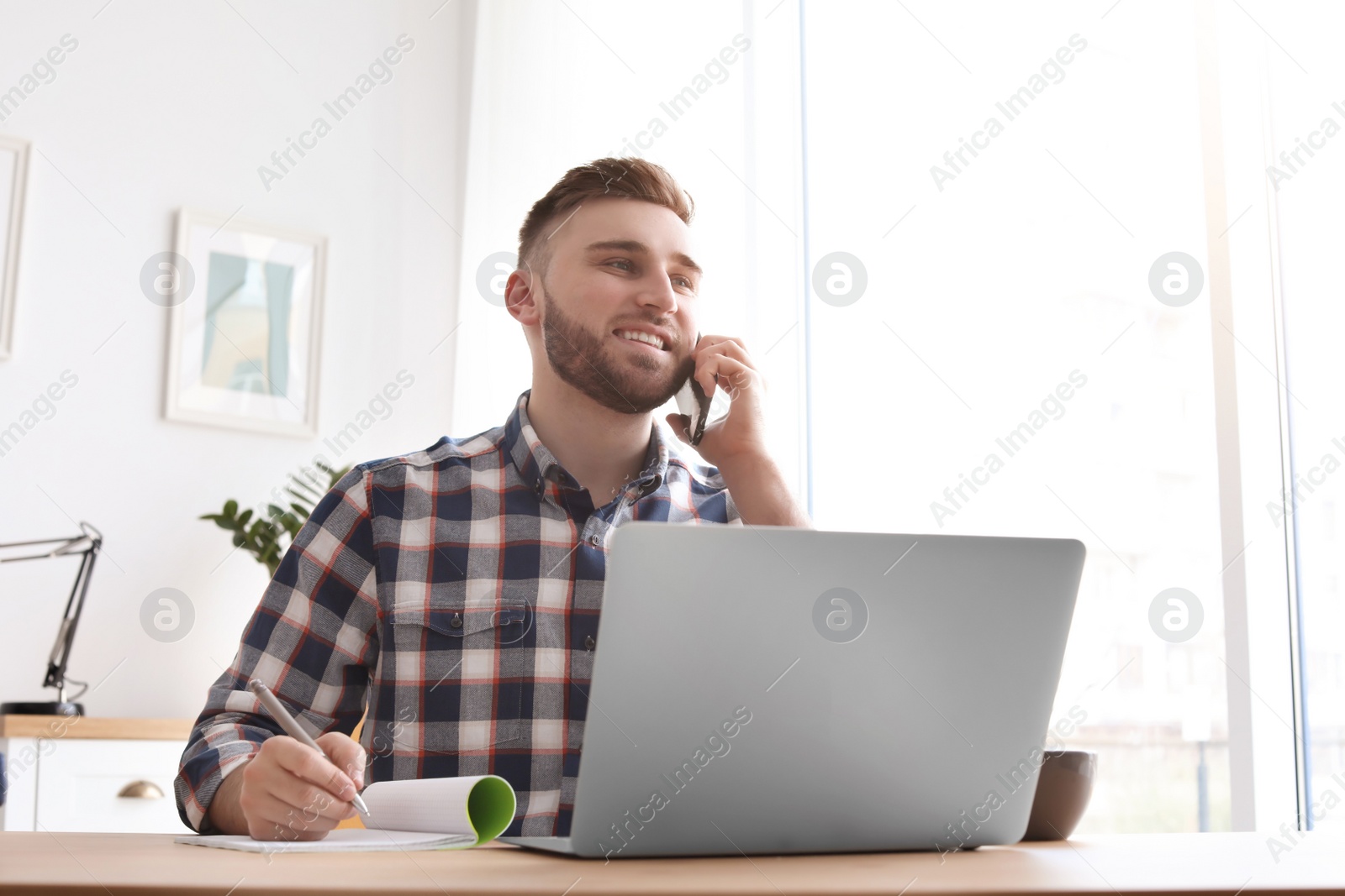 Photo of Young man talking on mobile phone while working with laptop at desk. Home office