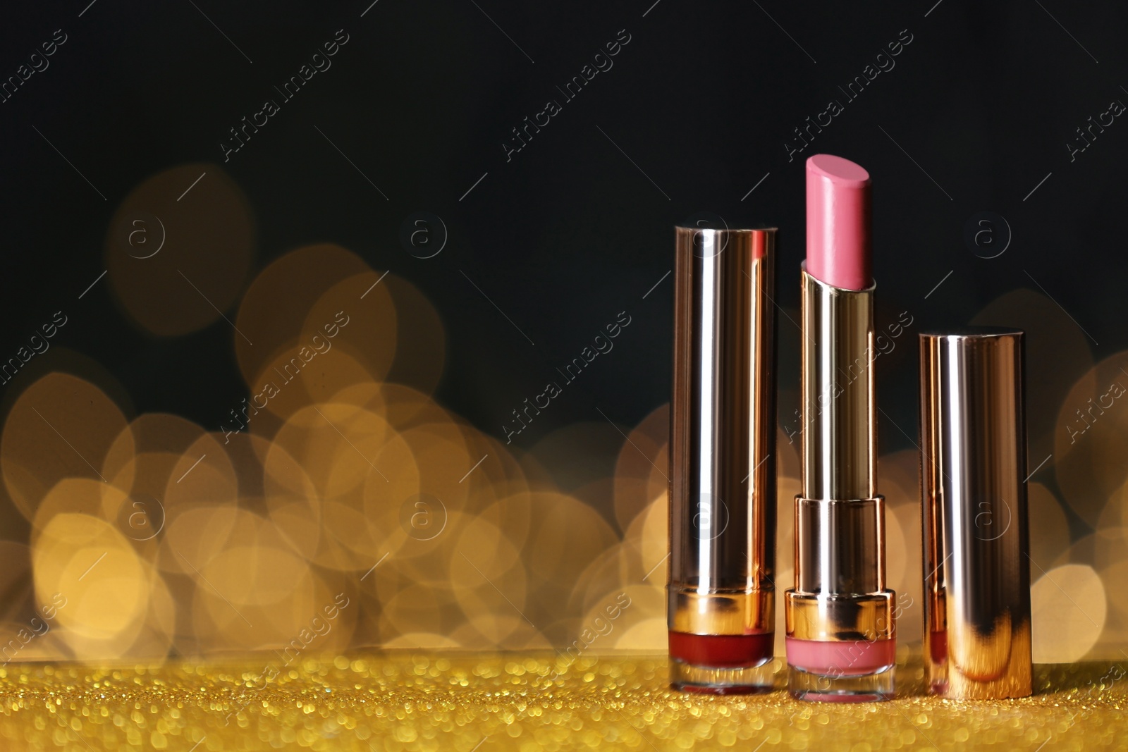 Photo of Bright lipsticks on table with gold glitter against blurred lights, space for text