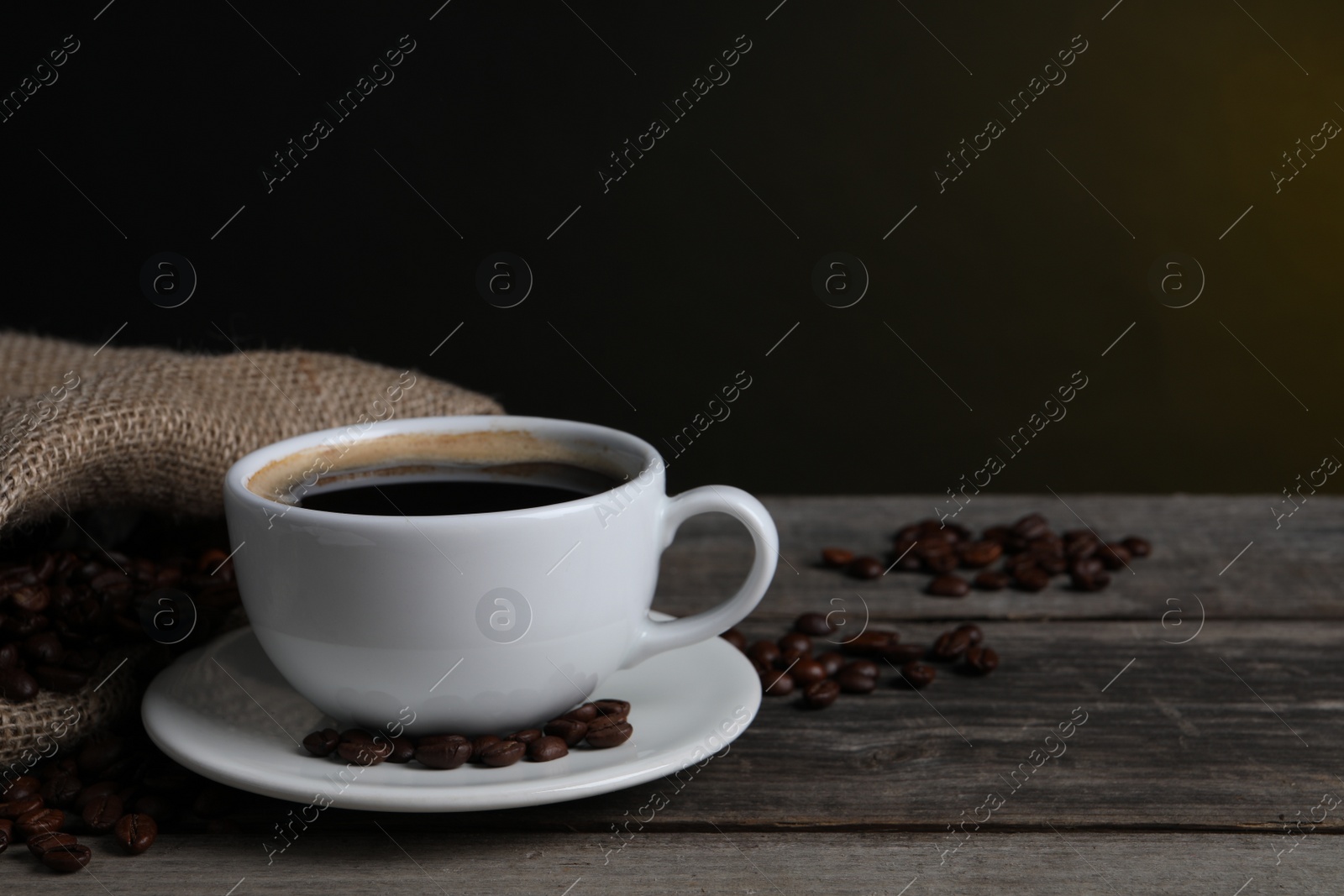 Photo of Cup of hot aromatic coffee and roasted beans on wooden table against dark background, space for text