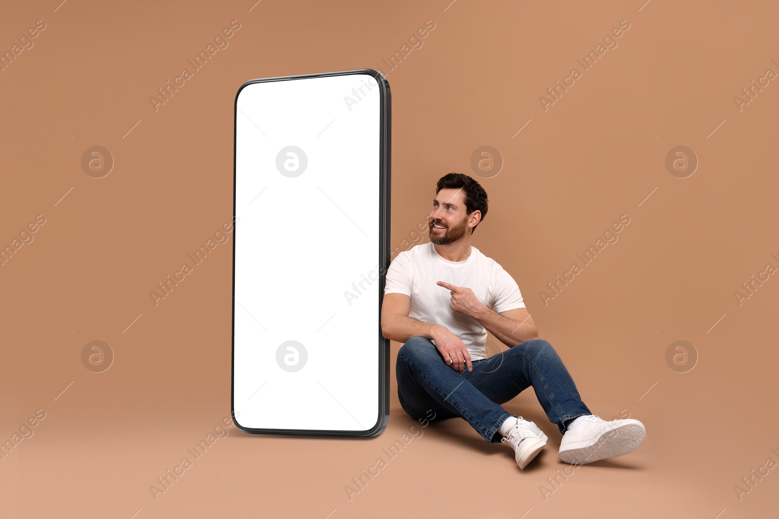 Image of Man sitting near huge mobile phone with empty screen on dark beige background. Mockup for design